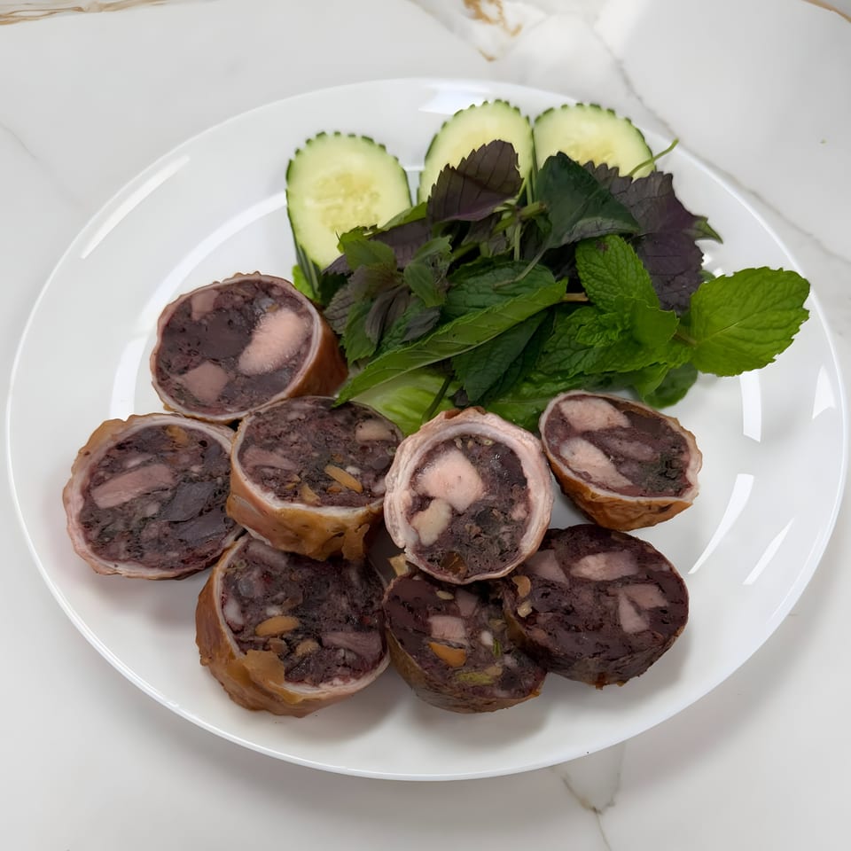 Sliced Vietnamese blood sausage (dồi tiết) with fresh mint, perilla, and cucumber on a white plate.