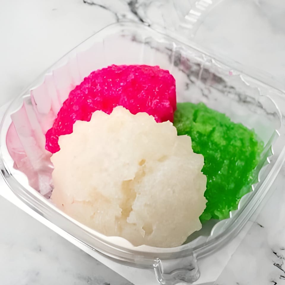 Three colorful Vietnamese steamed rice cakes, pink, white, and green, in a clear plastic container.