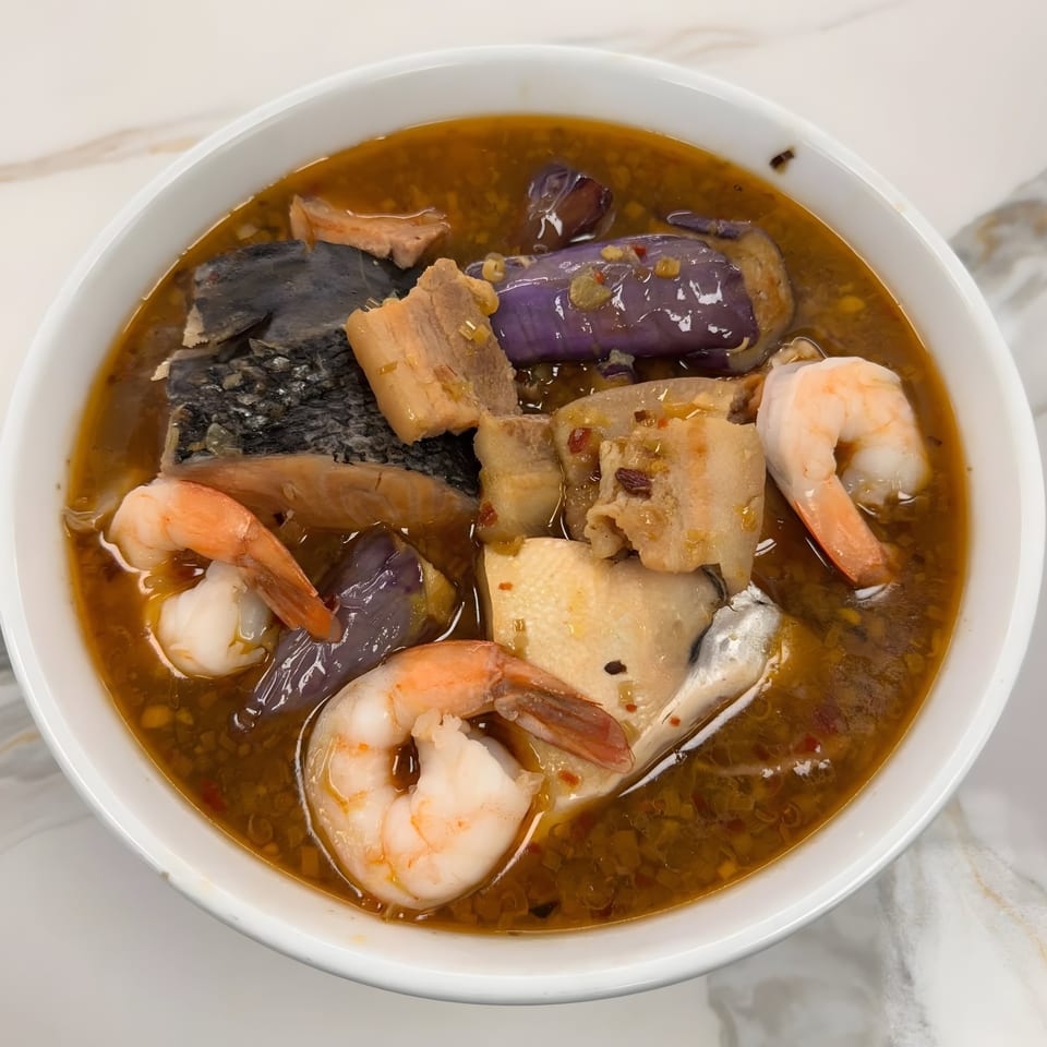 A bowl of Vietnamese canh chua with shrimp, fish, pork belly, and purple eggplant in a rich broth.