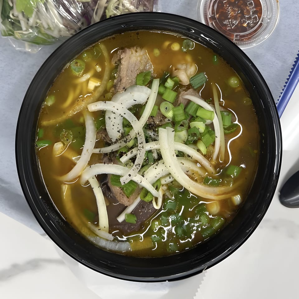 A close-up of a Vietnamese Bun Bo Hue soup in a black bowl, topped with onions and scallions.
